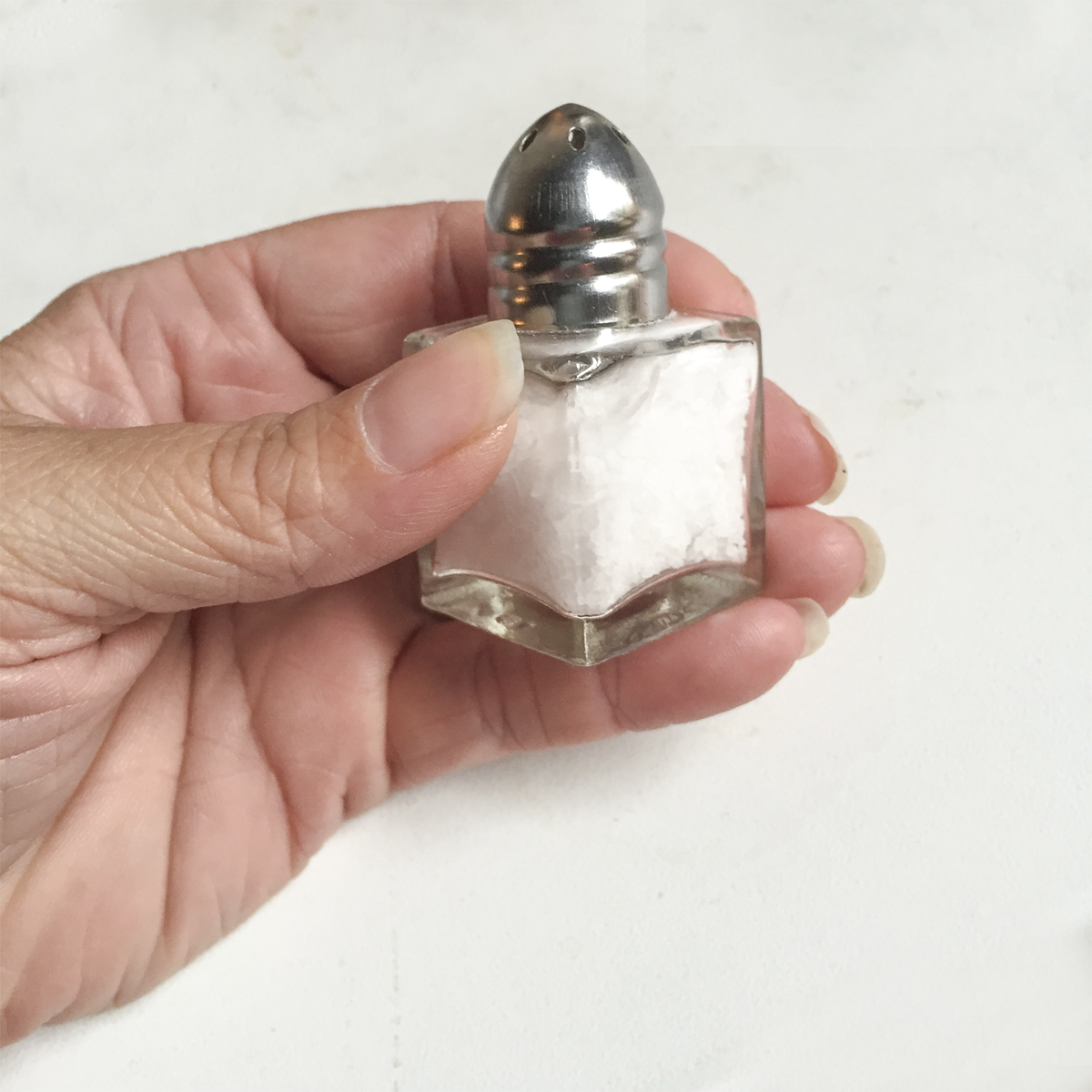 A photo of a small salt shaker. A woman's hand holds a small, cubic shaped salt shaker that has an approximate edge length of the width of 2 of her fingers.