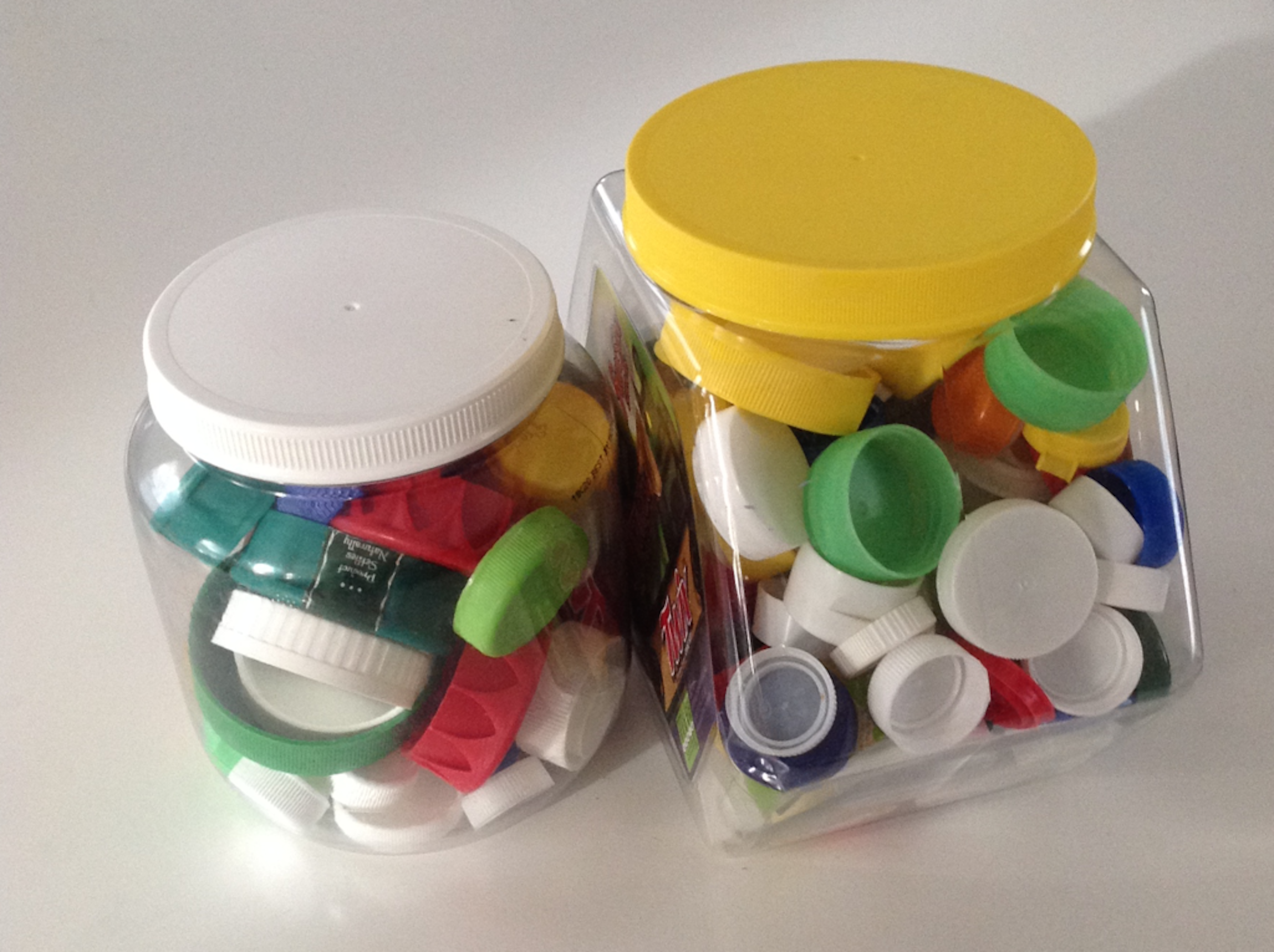 A photo of two clear, plastic containers containing plastic bottle caps of various sizes and colors. The colors of the plastic bottle caps are red, orange, yellow, green, blue, and white.